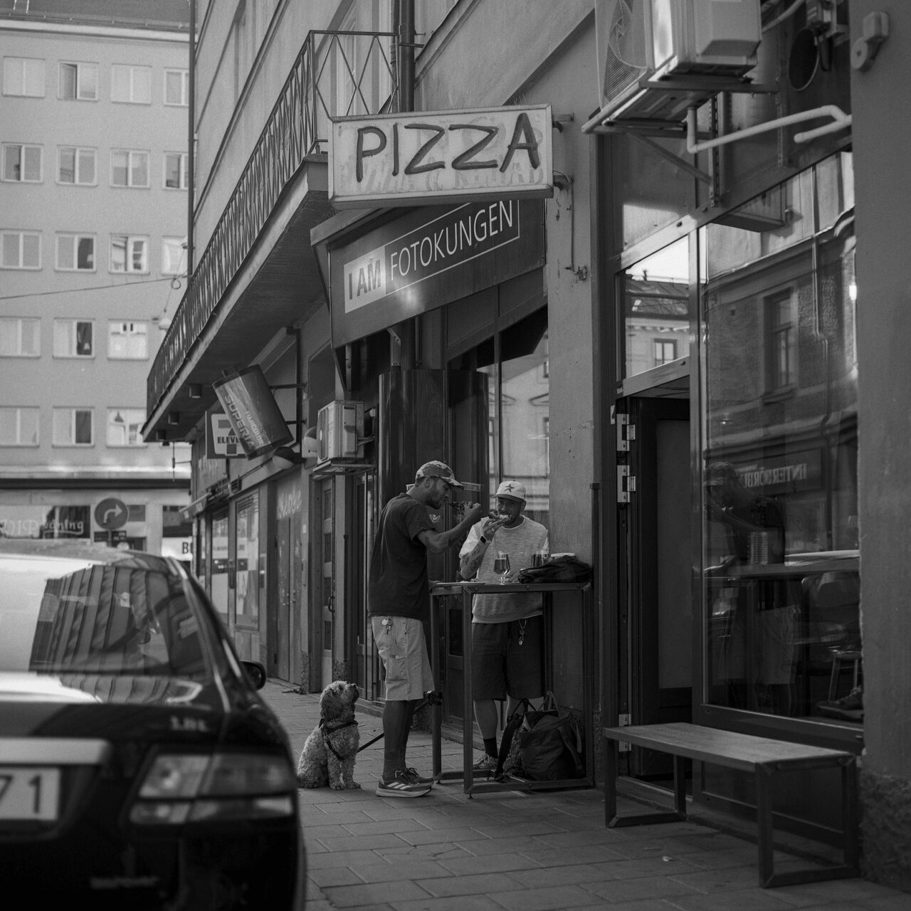 Two people eating a slice of pizza in the street outside a small restaurant.
